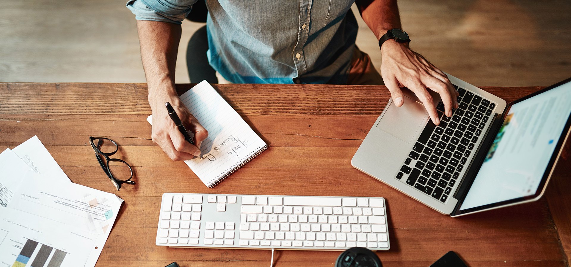 View from above of a desk with a laptop and a person holding paper and pencil