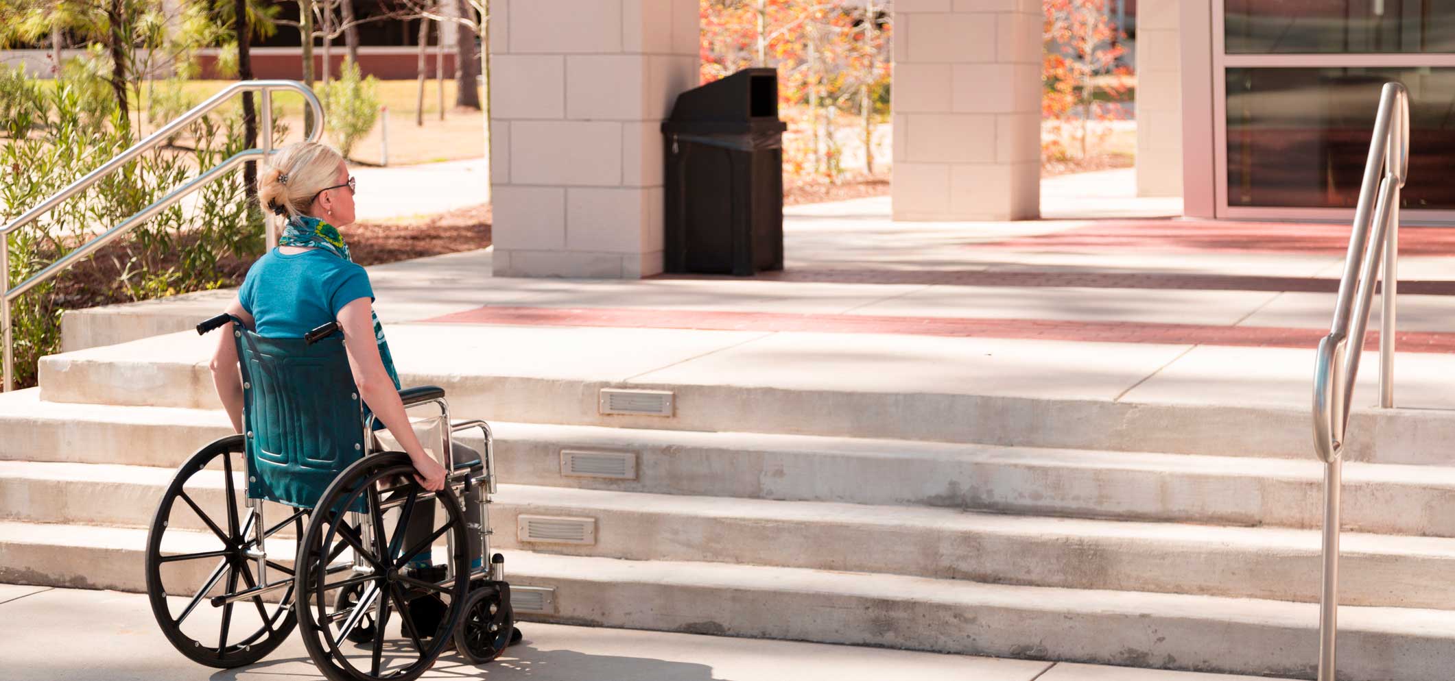 A women in a wheelchair at the bottom of some steps. The steps lead to the entrance of a building.