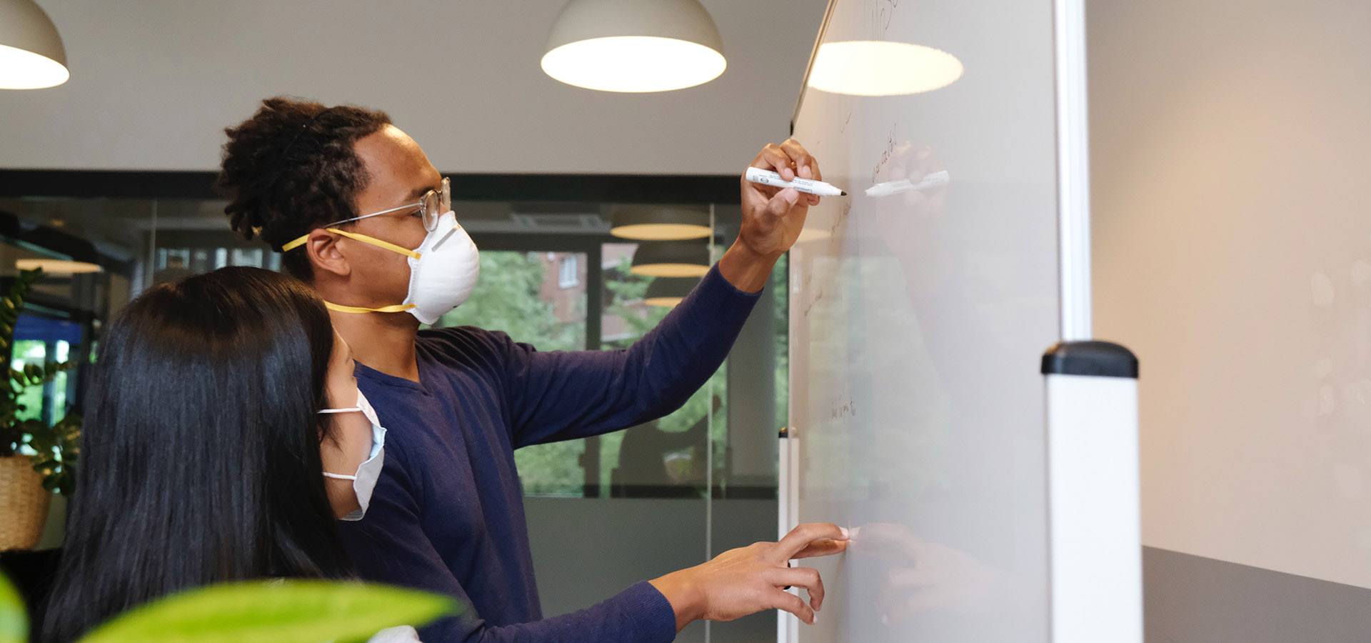 Two people wearing face masks stand at a white board